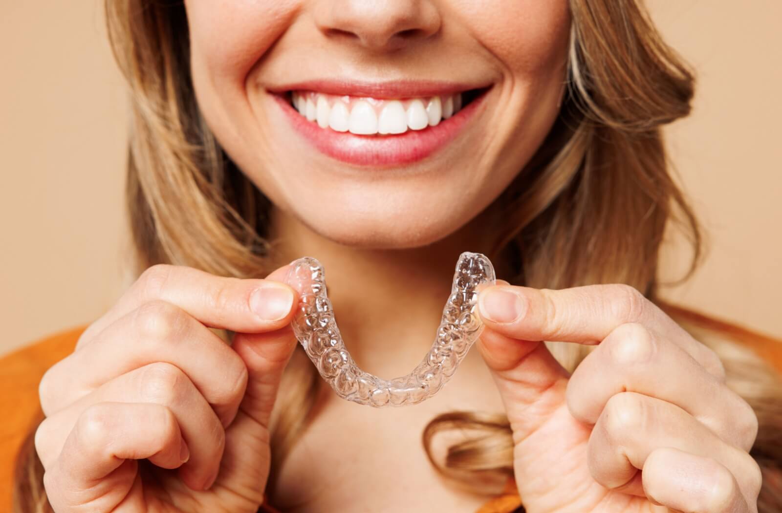 Close-up of a smiling person holding a clear Invisalign retainer near their mouth.