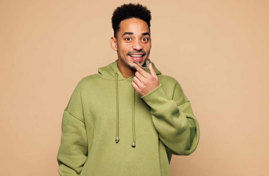 Smiling young adult in green hoodie holding clear aligners (Invisalign) to mouth against a pale orange background.