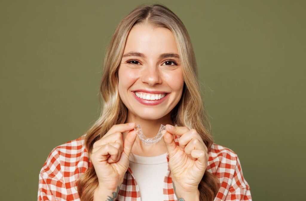 Person smiling and holding a clear orthodontic aligner with both hands against a plain background.