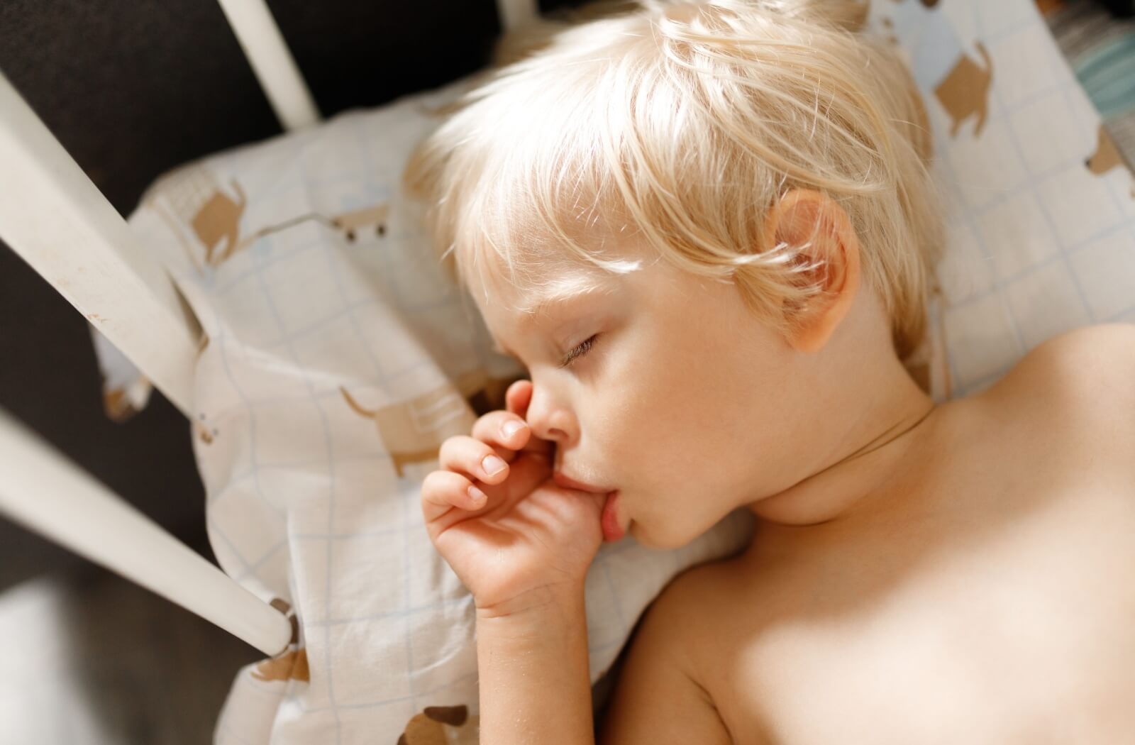 Small cute blonde child sleeping in his white crib and sucking his thumb finger. sucking reflex