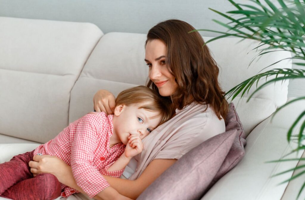 Baby girl is sucking finger lying on mothers chest mother on sofa at home interior. Smiling Mom and daughter in Pajamas lie hug resting together on couch
